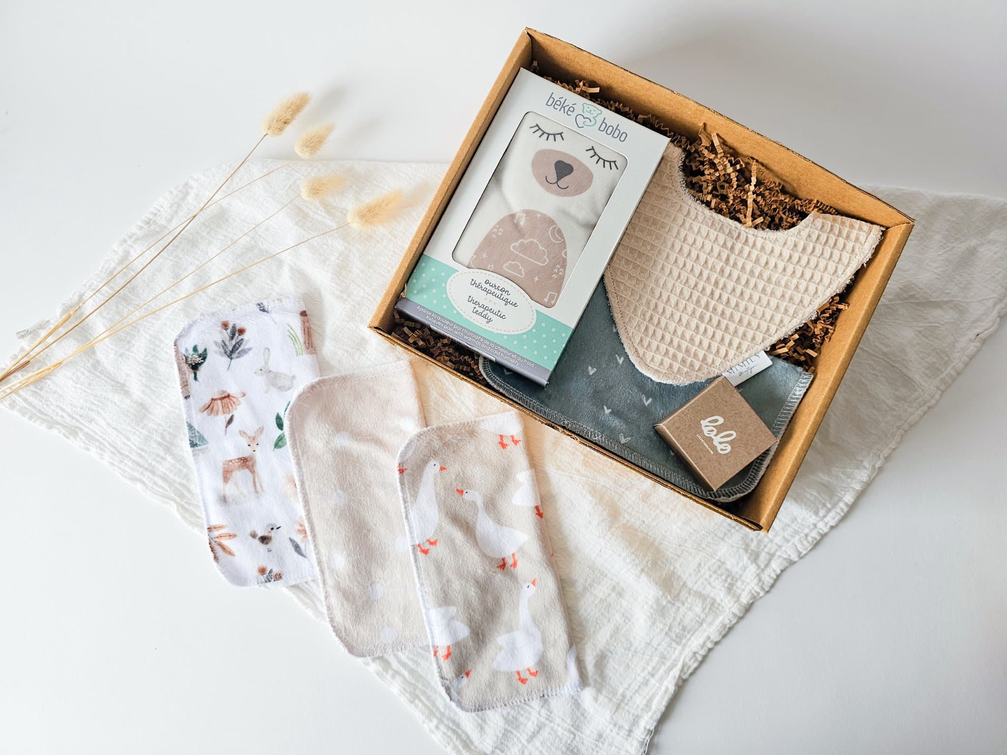 Boîte cadeau Petit trésor pour nouveau-né avec ourson chaud-froid Béké bobo, bavoir absorbant, débarbouillette douce et savon naturel, présentée sur couverture en mousseline, idée cadeau naissance pratique et unisexe.